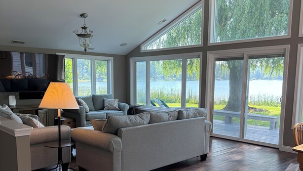 Ein geräumiges Wohnzimmer mit einem großen grauen Sofa, einem kleinen Tisch mit einer Lampe und einem Deckenventilator. Große Fenster bieten Blick auf einen See und Bäume.