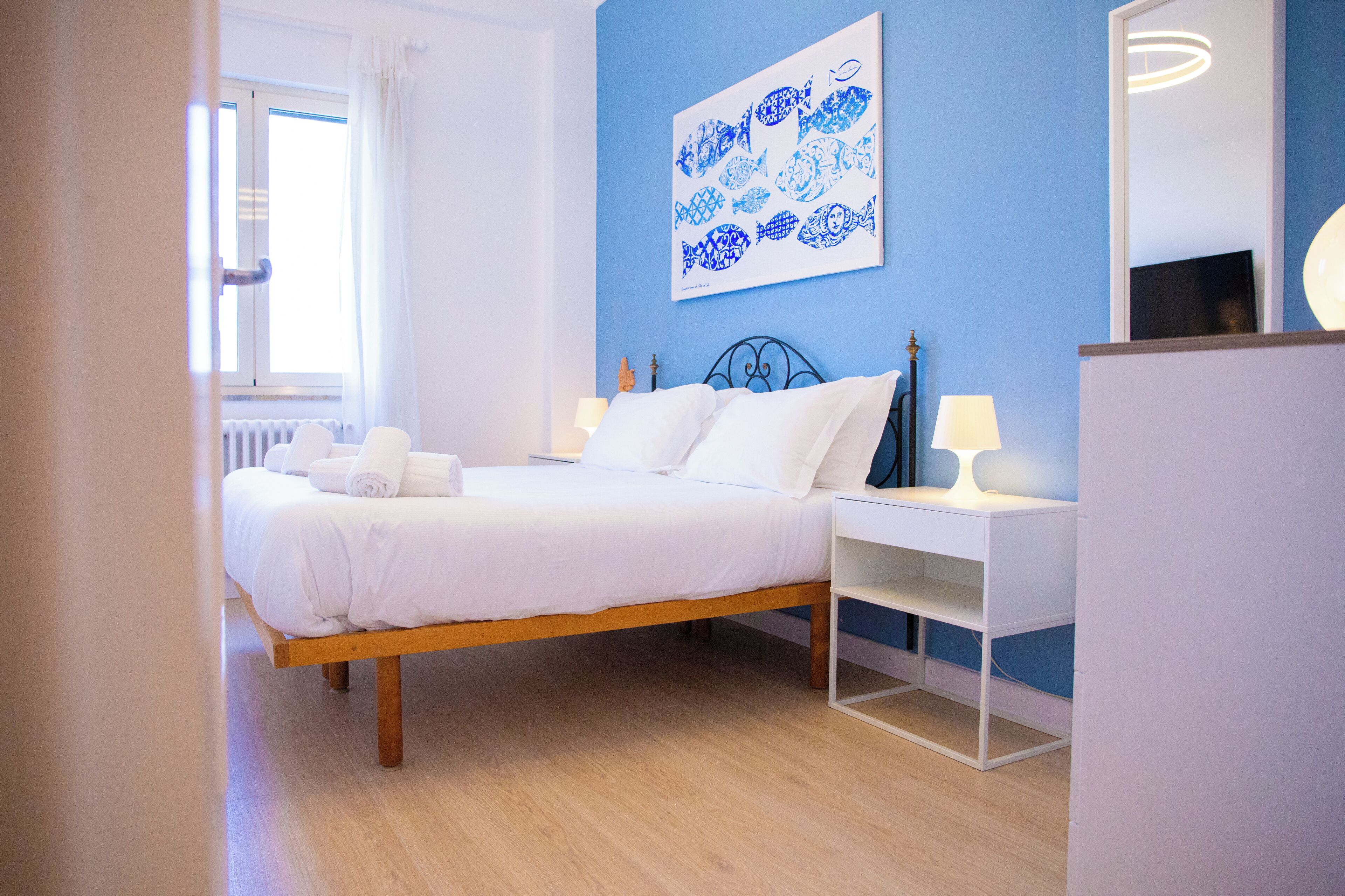 A bedroom with a wooden bed, white bedding, a blue wall, and a framed artwork.