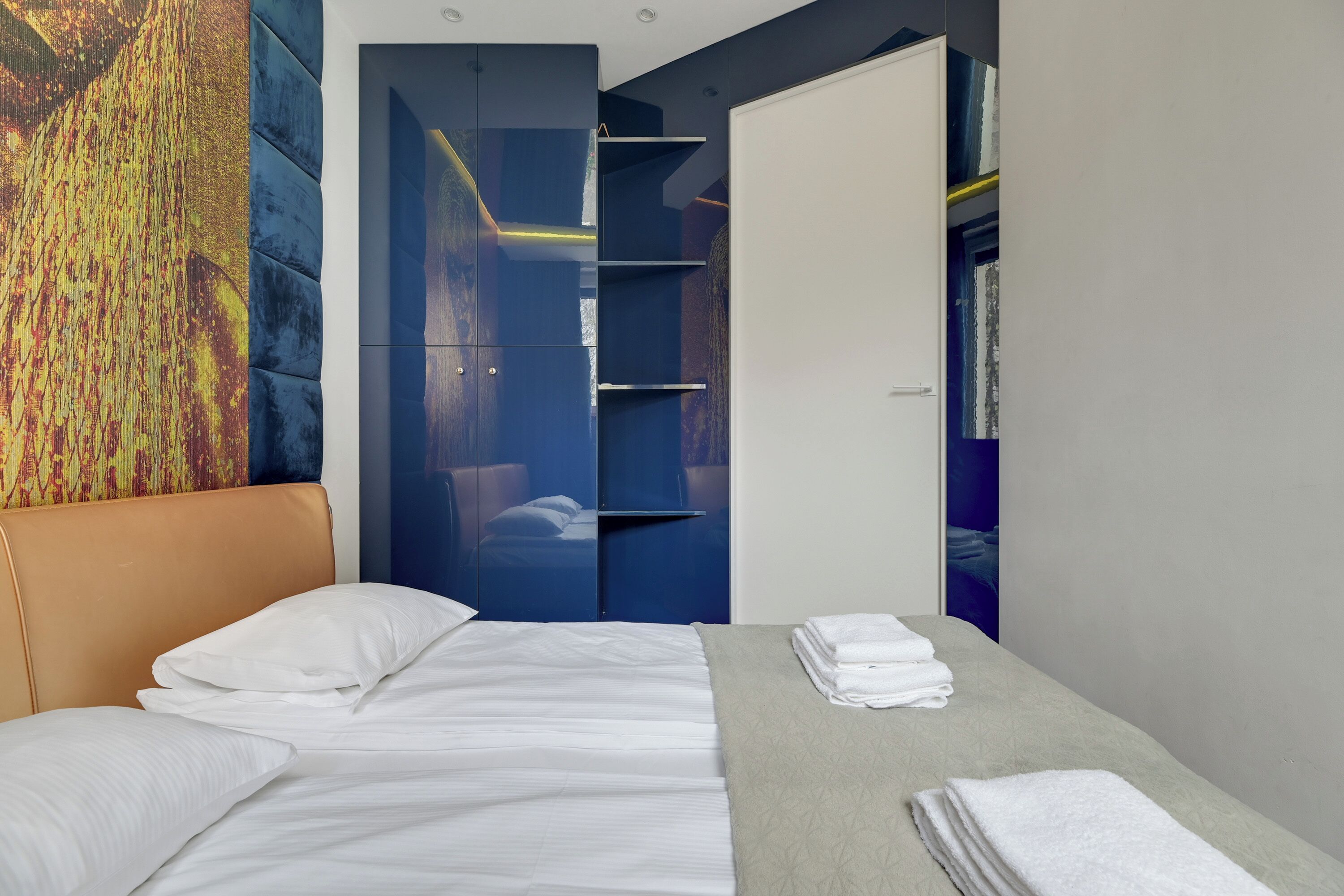 Ett modernt hotellrum med en säng, ett litet bord med handdukar, en garderob och en färgstark, abstrakt målning på väggen.