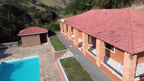 A poolside area with a swimming pool, a brick building with a red roof, and a view of hills in the background. - Chácara Paraíso de Itamonte (Itanhandu)
