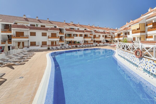 Une piscine avec des chaises longues et une clôture blanche, entourée d’immeubles à plusieurs étages dotés de balcons.
