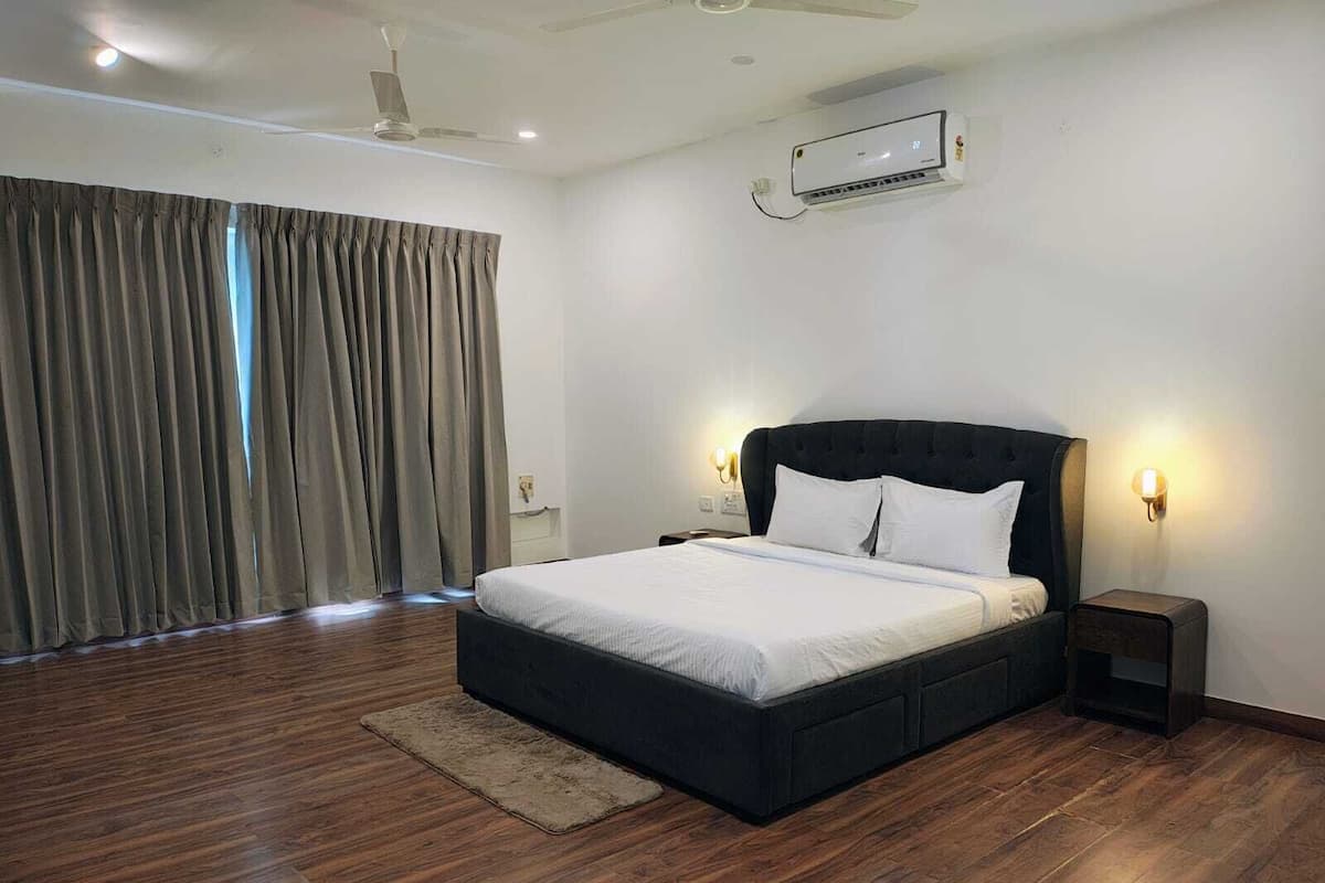 Une chambre d’hôtel moderne avec un grand lit, un plancher en bois, un ventilateur de plafond, un appareil de climatisation et une vue sur l’extérieur à travers les rideaux.
