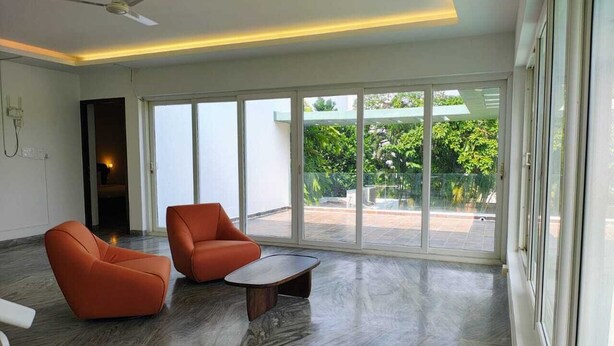 A spacious room with a large sliding glass door leading to a balcony, a ceiling fan, and a modern seating area with two orange chairs and a small table. - Truliv Villa Nivara (Tirupporur)