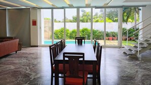 A spacious indoor area with a long table and chairs, large windows, and a staircase leading to an outdoor area with plants. - Truliv Villa Nivara (Tirupporur)