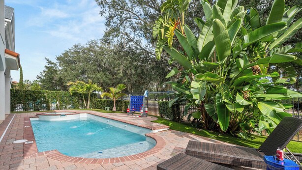 A swimming pool with a brick border, surrounded by lush greenery and a fence. - Reunion villa w/ pool & cinema room (Kissimmee)