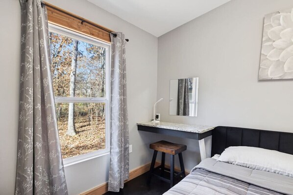 Un dormitorio con cama, mesita de noche, ventana con cortinas, espejo y vista a árboles.