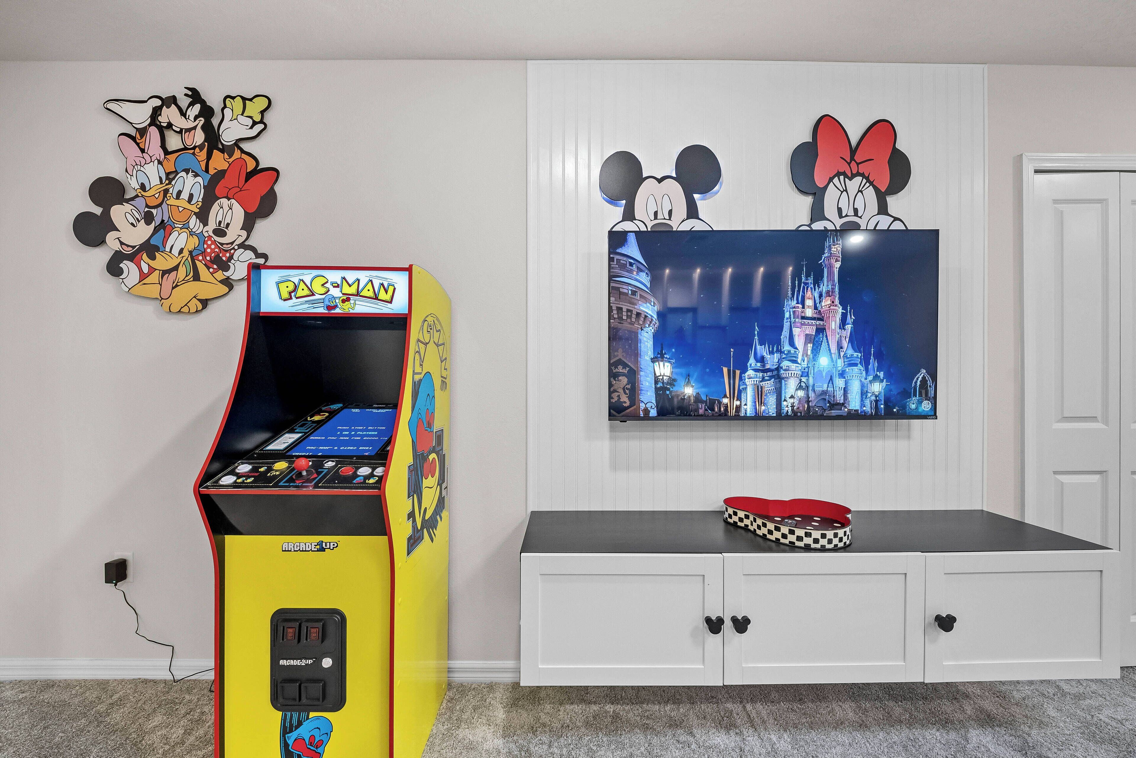 A room with a Pac-Man arcade machine, a television displaying a castle scene, and a white cabinet with a Mickey Mouse cutout.