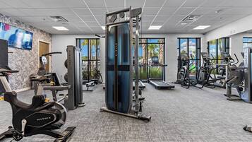 A modern gym with cardio machines, weight training equipment, and large windows offering views of palm trees.
