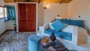 A room with a wooden door, a white sofa with blue cushions, a small round table with a remote control, and a potted plant. - Escale Nature near Marrakech | Private Bungalows, Pool, Terrace & Hammam (Marrakech)