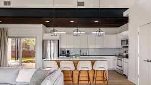 A modern kitchen with white cabinets, a central island with bar stools, and a dining area with a grey sofa. - Modern Hill Country Home ~ Sunset Views and BBQ (Austin)
