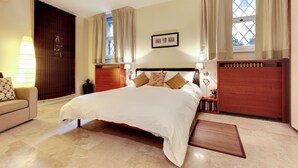 Habitación de hotel con una cama grande, una mesita de noche y un cuadro colgado en la pared. Se ve una ventana con cortinas y una puerta de madera.