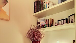 Una habitación con un sofá blanco, una estantería con libros y objetos decorativos, un jarrón con flores secas y un cuadro en la pared.