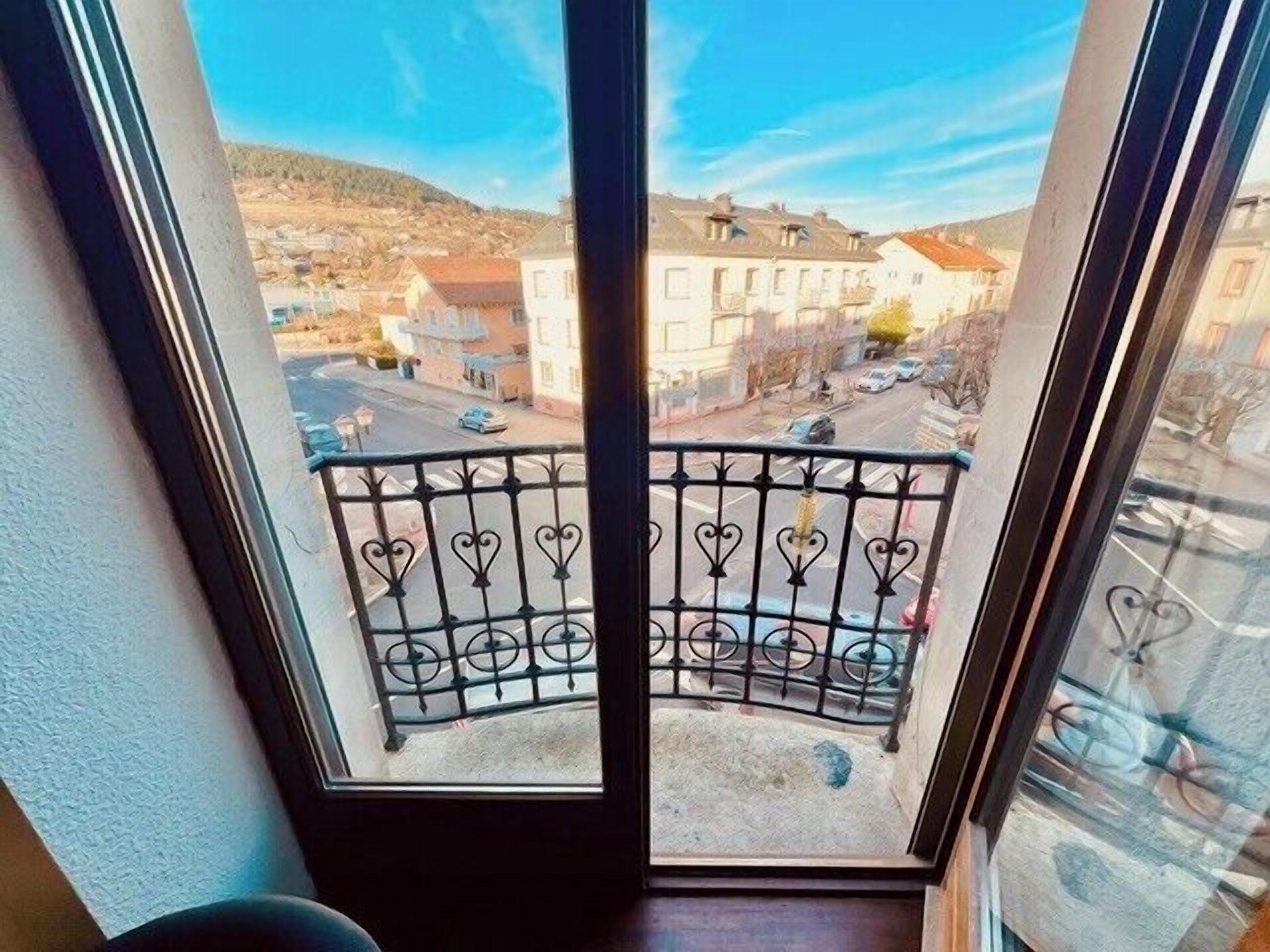 A balcony with a wrought iron railing overlooking a street with buildings and a hilly landscape.