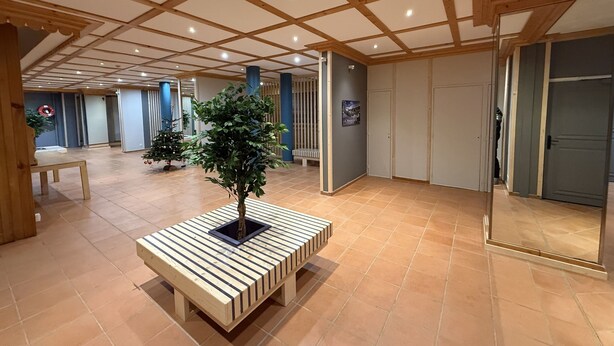 A spacious lobby with a wooden ceiling, a central wooden bench with a small tree, and a Christmas tree in the background. - Bright studio in Risoul, ski on foot and comfort (Risoul)