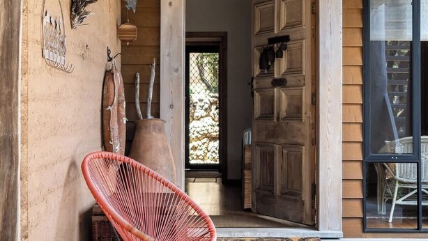 Une façade en bois avec un porche abrité, une chaise en osier rouge et un intérieur rustique visible à travers une porte ouverte.
