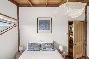 Ein Schlafzimmer mit einem Bett, zwei Nachttischlampen, einem gerahmten Bild an der Wand, einem Fenster und einer Tür zu einem weiteren Raum.