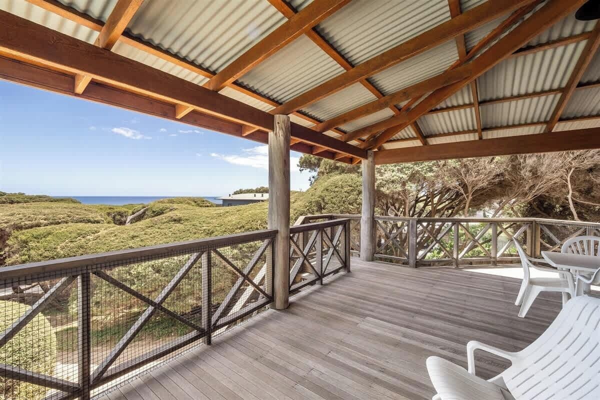 A covered outdoor deck with a view of the ocean, wooden railings, and white plastic chairs.