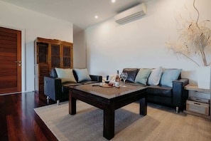 A living room with a dark wooden coffee table, two black sofas with light-colored cushions, a white air conditioning unit on the wall, and a decorative plant on the right.