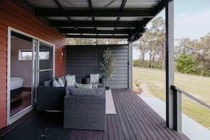 A covered outdoor seating area with a sofa and a small table, a potted plant, and a view of a grassy area.