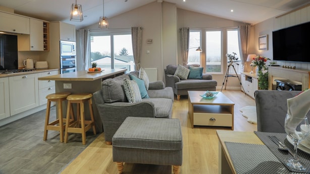 A modern living room with a grey sofa, ottoman, and wooden coffee table. A kitchen with white cabinets and a built-in oven is visible. - Argyll View Lodge (Wemyss Bay)