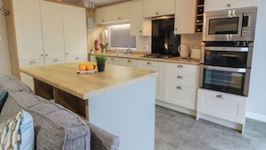 A modern kitchen with white cabinets, a wooden island, stainless steel appliances, and a grey sofa in the living area. - Argyll View Lodge (Wemyss Bay)