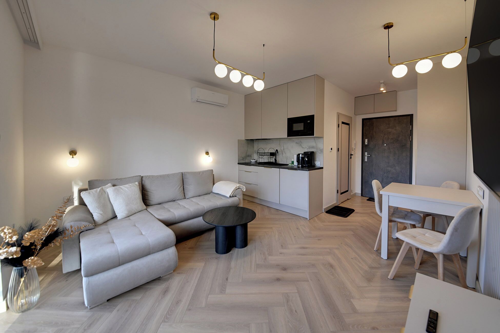 Een moderne woonkamer met een grijze modulaire bank, een eettafel met stoelen, een keuken met een magnetron en een zwarte deur.