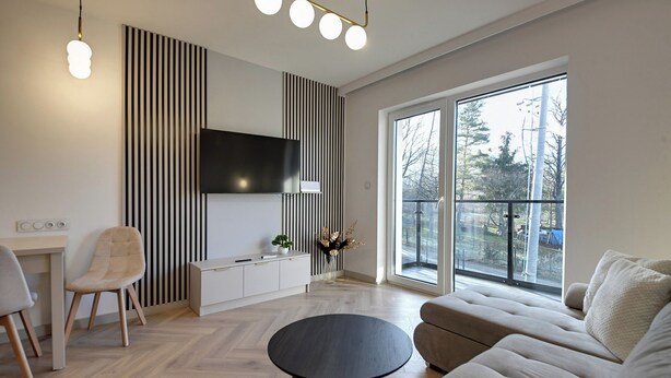 Ein modernes Wohnzimmer mit großem Fenster, einem an der Wand befestigten Flachbildfernseher, einem Sofa, einem Couchtisch und einem Sessel.