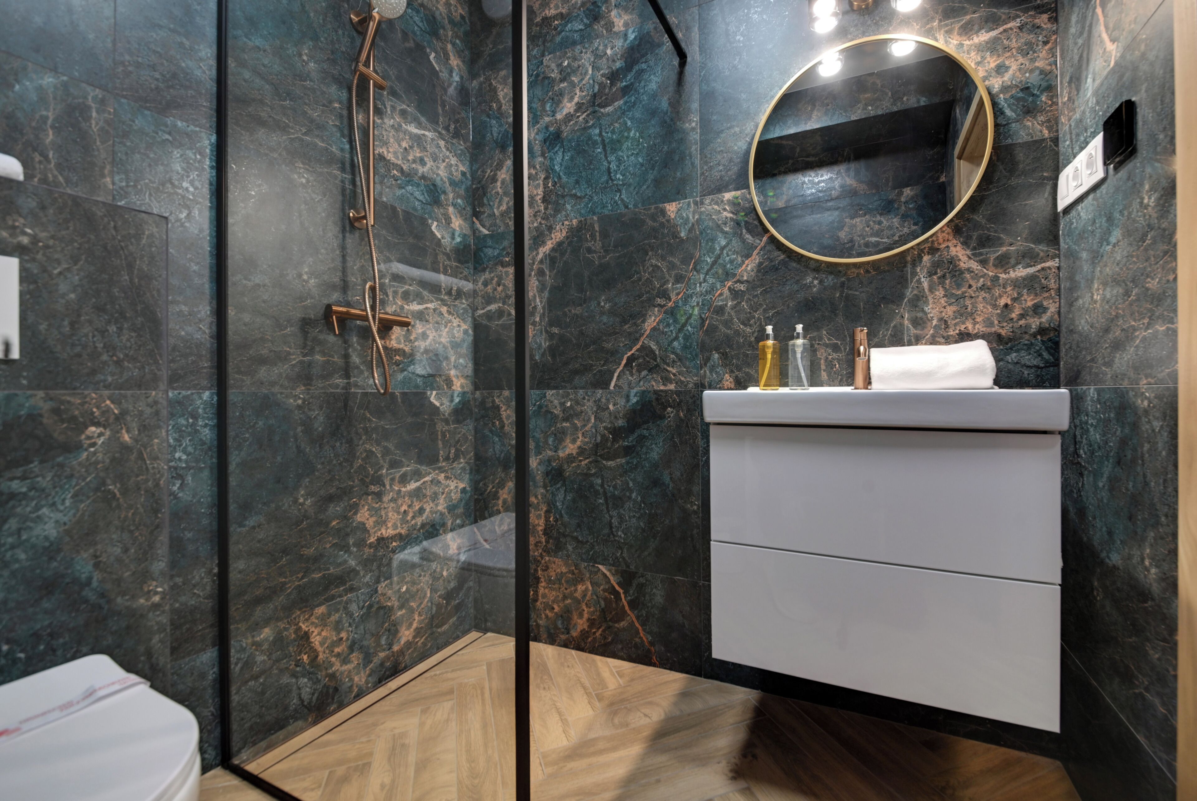 Een moderne badkamer met een douchecabine van glas, een marmeren wand, een ronde spiegel en een witte wastafelmeubel.