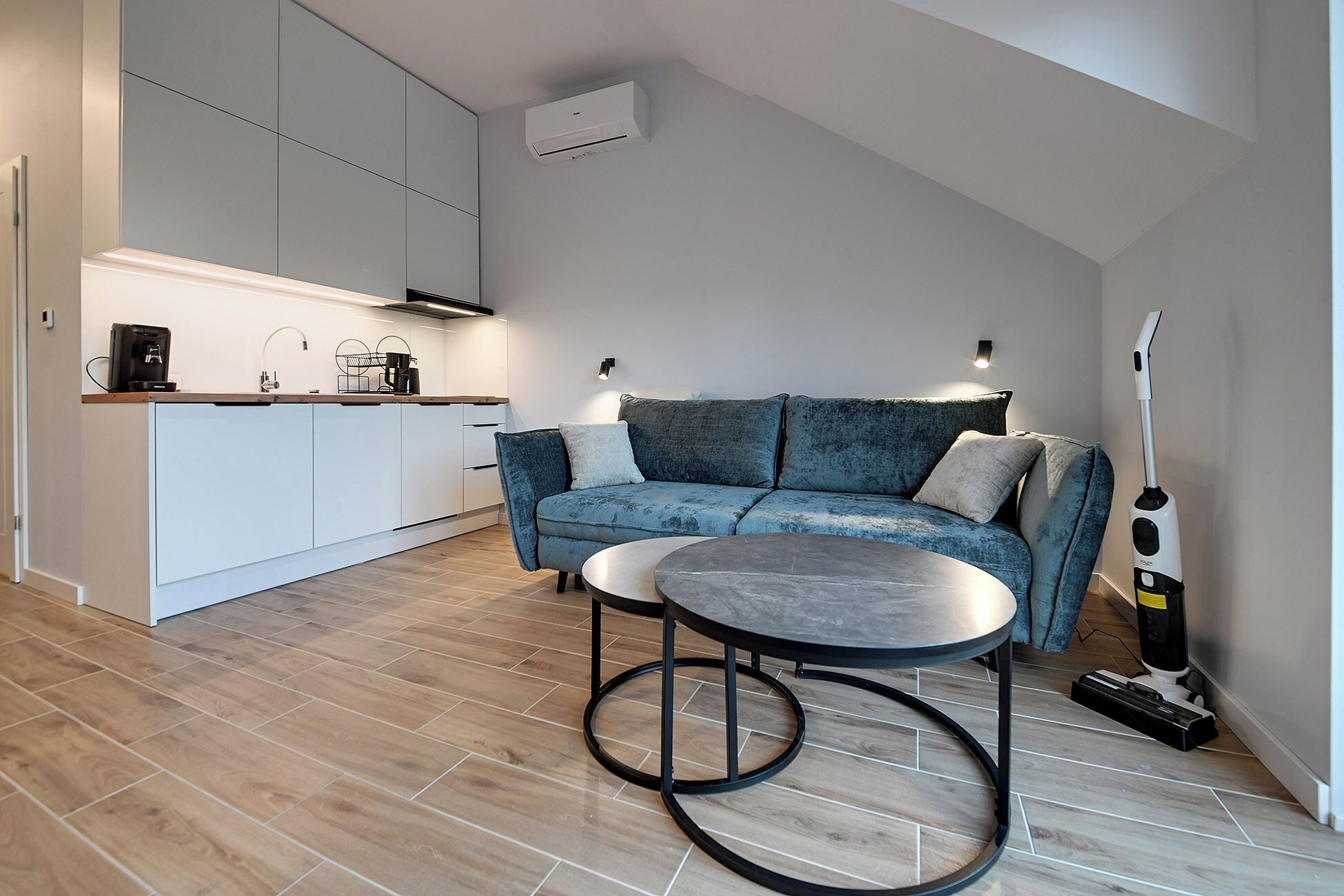 Un espacio de estar moderno con cocina, un sofá azul, una mesa de centro redonda y un aspirador.
