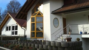 Une maison à deux étages dotée d’une grande baie vitrée, d’un escalier menant à l’étage supérieur et d’un petit balcon agrémenté de plantes en pot.