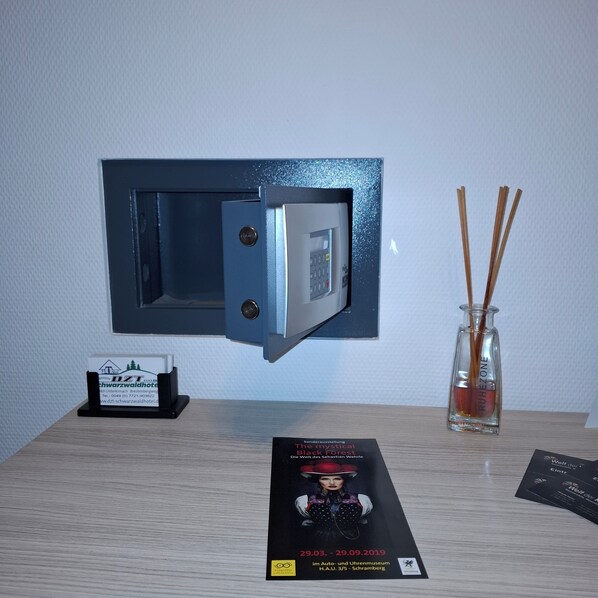 Une pièce comprenant un coffre-fort fixé au mur, une petite table avec une brochure, un vase en verre contenant des tiges de roseau et un présentoir à cartes de visite.