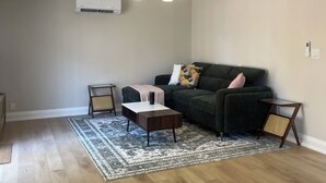 A living room with a dark green sofa, a coffee table, and a patterned rug. There is a ceiling light fixture and an air conditioning unit on the wall. - 1 Bed, 1 Bath Private Bungalow in downtown Thorold (Thorold)