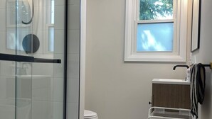 A modern bathroom with a glass shower enclosure, a toilet, a sink with a wooden vanity, and a window providing natural light. - 1 Bed, 1 Bath Private Bungalow in downtown Thorold (Thorold)
