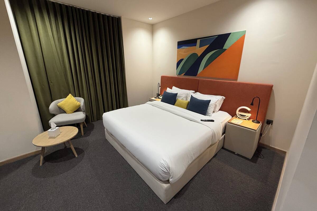 Hotelkamer met een groot bed, nachtkastje met een lamp, stoel met kussen, klein rond tafeltje met zakdoosje en een kleurrijk abstract schilderij aan de muur.