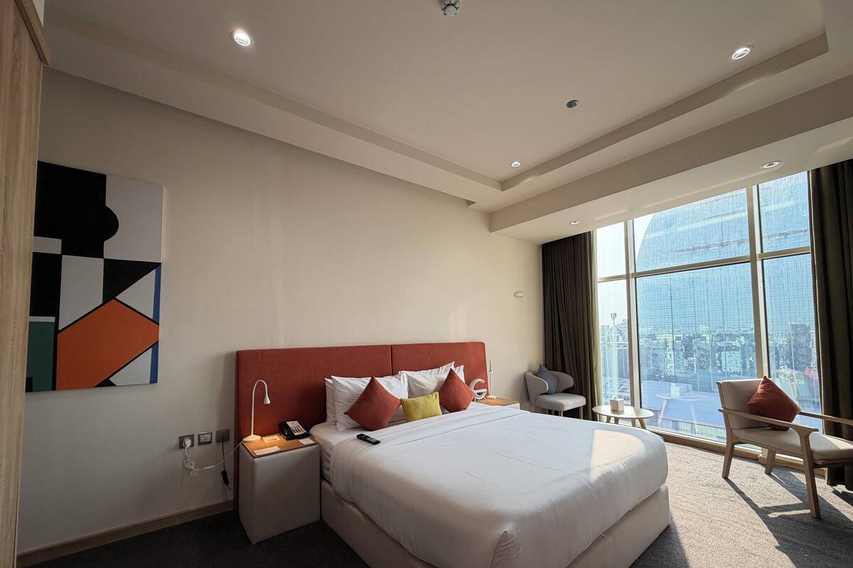 Een hotelkamer met een groot bed, een bureau, een stoel en uitzicht op de stad door grote ramen.