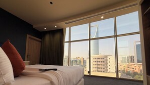 Habitación de hotel con un ventanal grande que ofrece vista a la ciudad, una cama con ropa blanca, un control remoto sobre la cama y una mesa de madera con un menú.