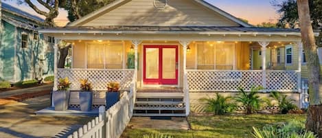 A single-story house with a red door, a porch with string lights, and a white picket fence.