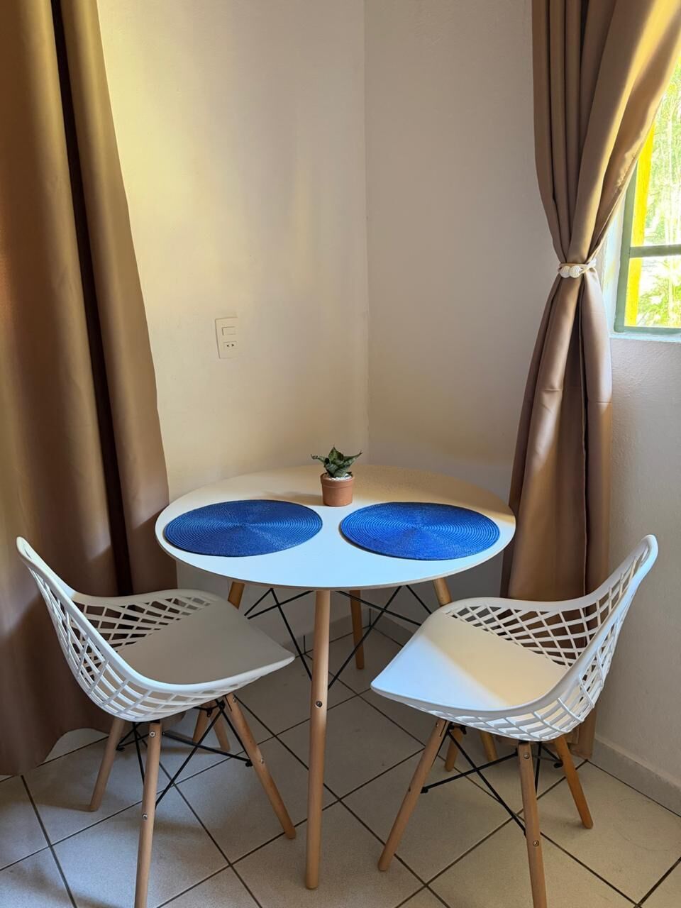 Quarto de canto com uma mesa redonda, duas cadeiras brancas, um vaso de planta pequeno e cortinas bege.