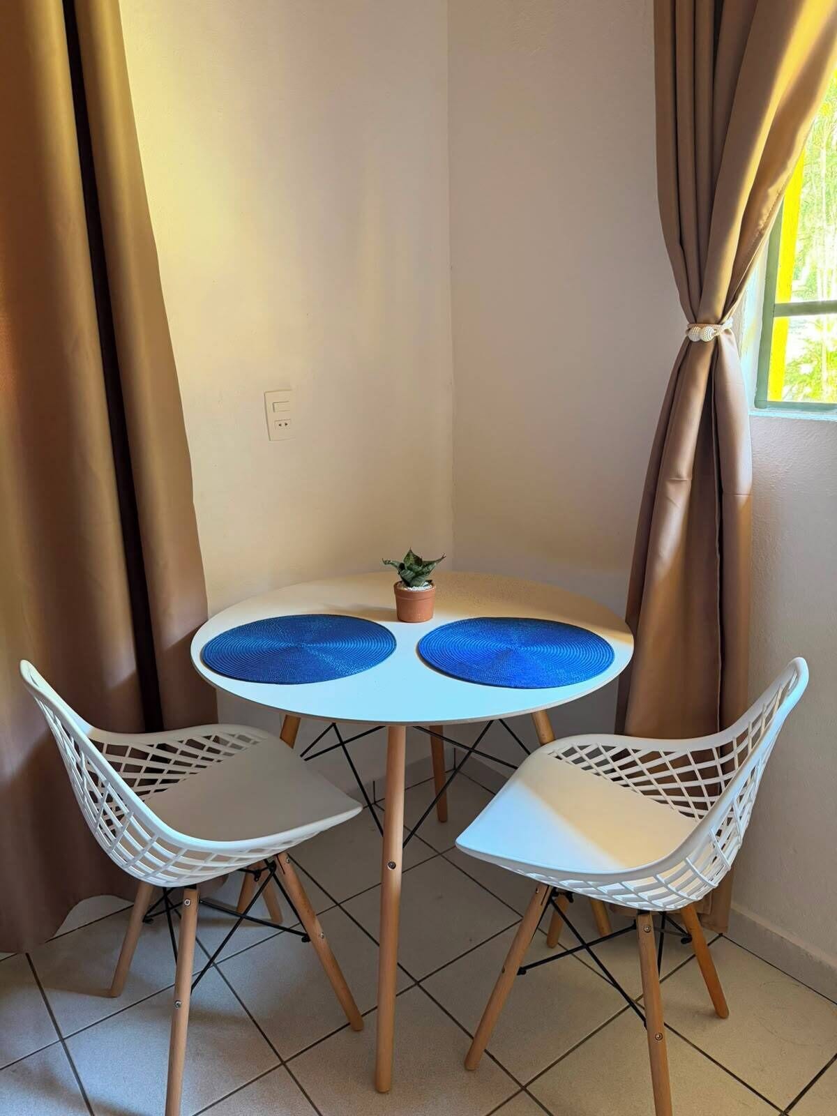 A small round table with two white chairs, a potted plant, and curtains.