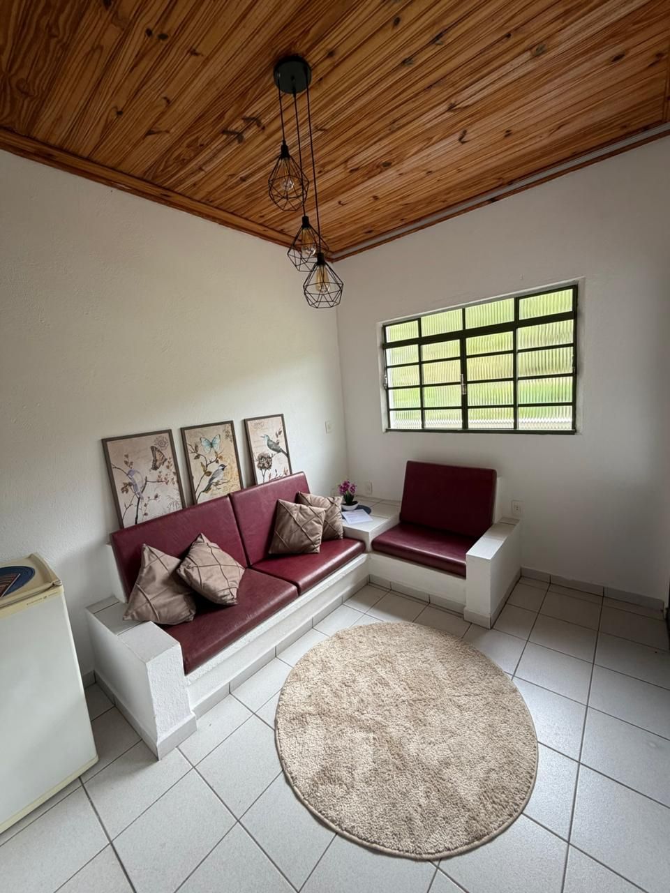 A living room with a wooden ceiling, a red sofa, a white sofa, a round rug, and a window with a grid pattern.