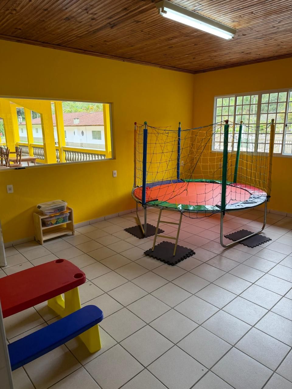 Une pièce avec un trampoline, des murs jaunes, un plafond en bois et une fenêtre donnant sur les bâtiments à l’extérieur.