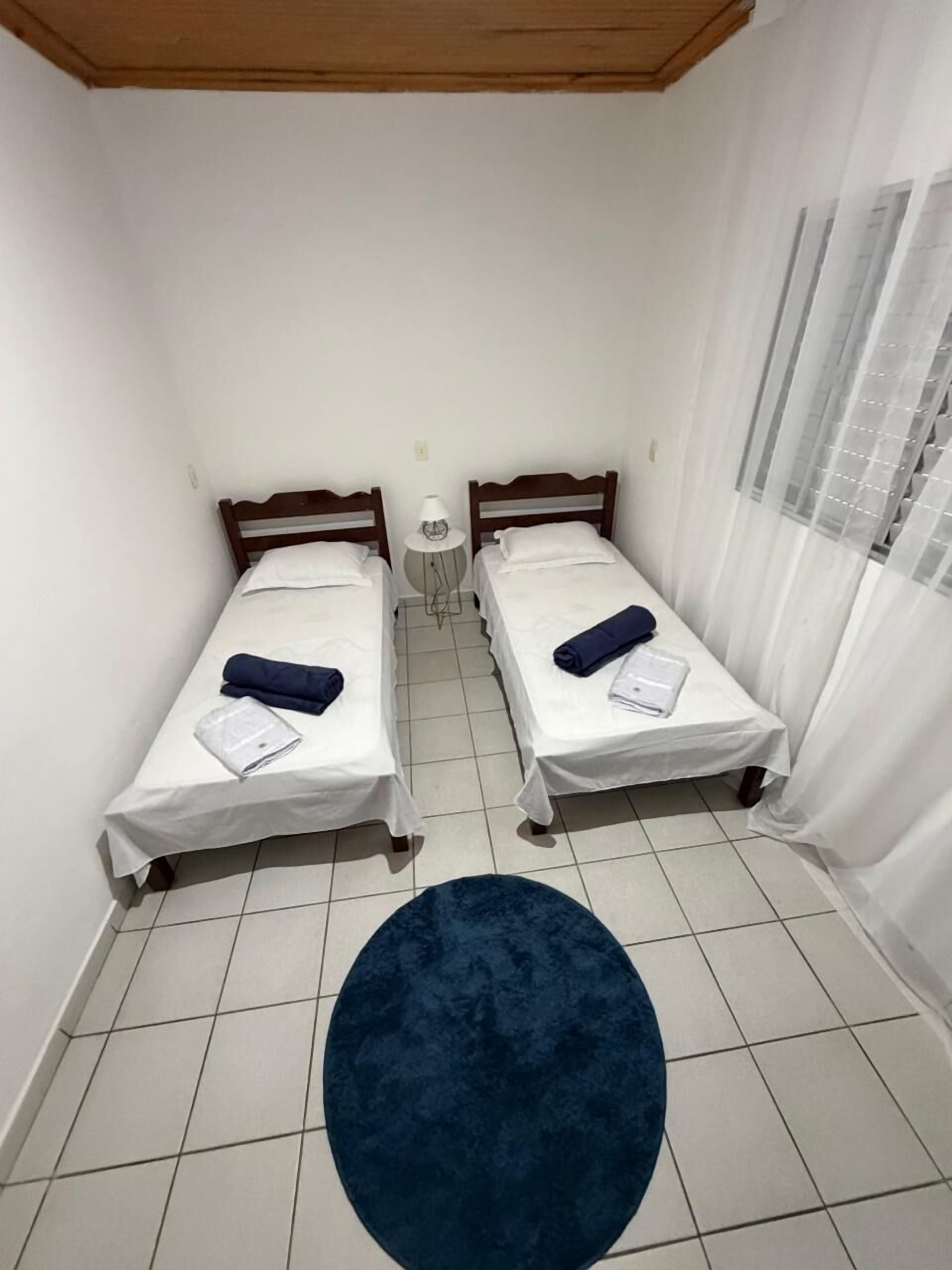 Una habitació petita i neta amb dos llits individuals, una catifa blava rodona i una finestra amb cortines transparents.
