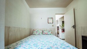 A bedroom with a bed, floral bedding, a mirror on the wall, and a door leading to another room. - Apartamento 350m da Praia do Bessa em Joao Pessoa (João Pessoa)