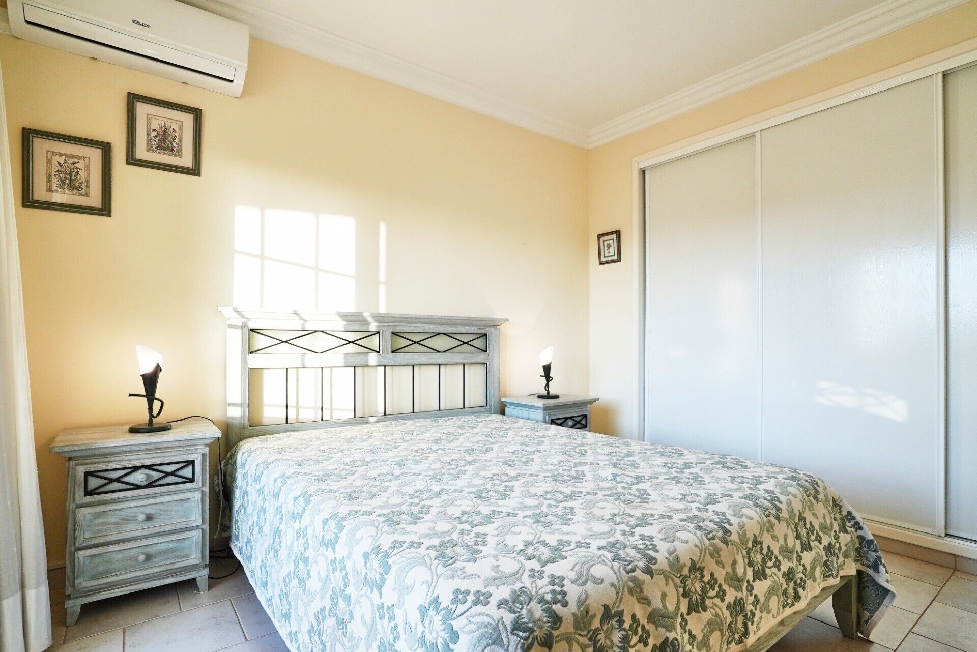 A bedroom with a bed, two bedside tables, a nightstand, a wardrobe, and an air conditioning unit.