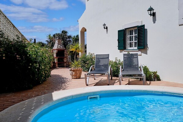 Une piscine avec deux chaises longues, une cuisine extérieure en pierre et un bâtiment blanc aux volets verts.