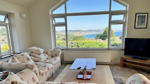 Un salon avec un canapé, une table basse et un téléviseur. De grandes fenêtres offrent une vue sur la mer et les collines.