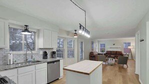 Cocina moderna con armarios blancos, lavavajillas integrado y una isla central. Hay un comedor con mesa y sillas, y una sala de estar con sofá y mesa de centro.