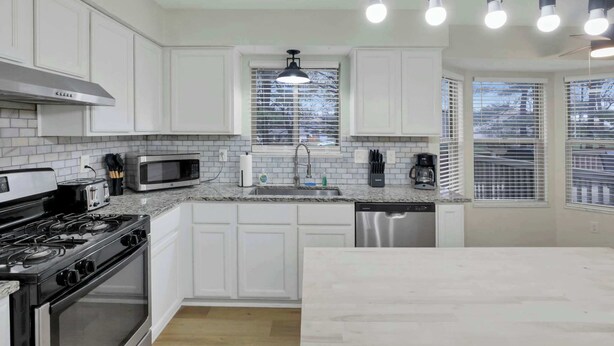 Cocina moderna con gabinetes blancos, estufa de acero inoxidable, microondas y lavavajillas. Cuatro lámparas colgantes sobre el fregadero y una ventana con persianas.