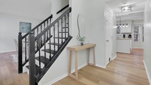 Un interior moderno con una escalera de madera, una consola con un florero y una zona de cocina al fondo.