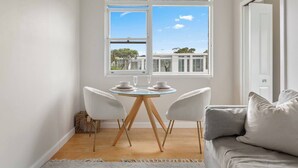 A modern room with a dining table set for two, a sofa, and a window with a view of buildings and trees. - The Little Lawrence Sunny Village Nook by the Sea (Freshwater)
